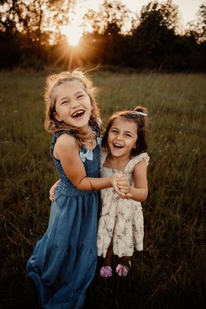 Two young girls in dresses laugh and embrace joyfully in a sunlit green field at sunset.