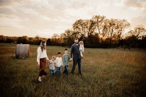 A family of six stands together in an open field at golden hour with a hay bale in the background.