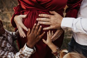 Multiple hands of family members lovingly touch a pregnant woman's belly in a red maternity dress.