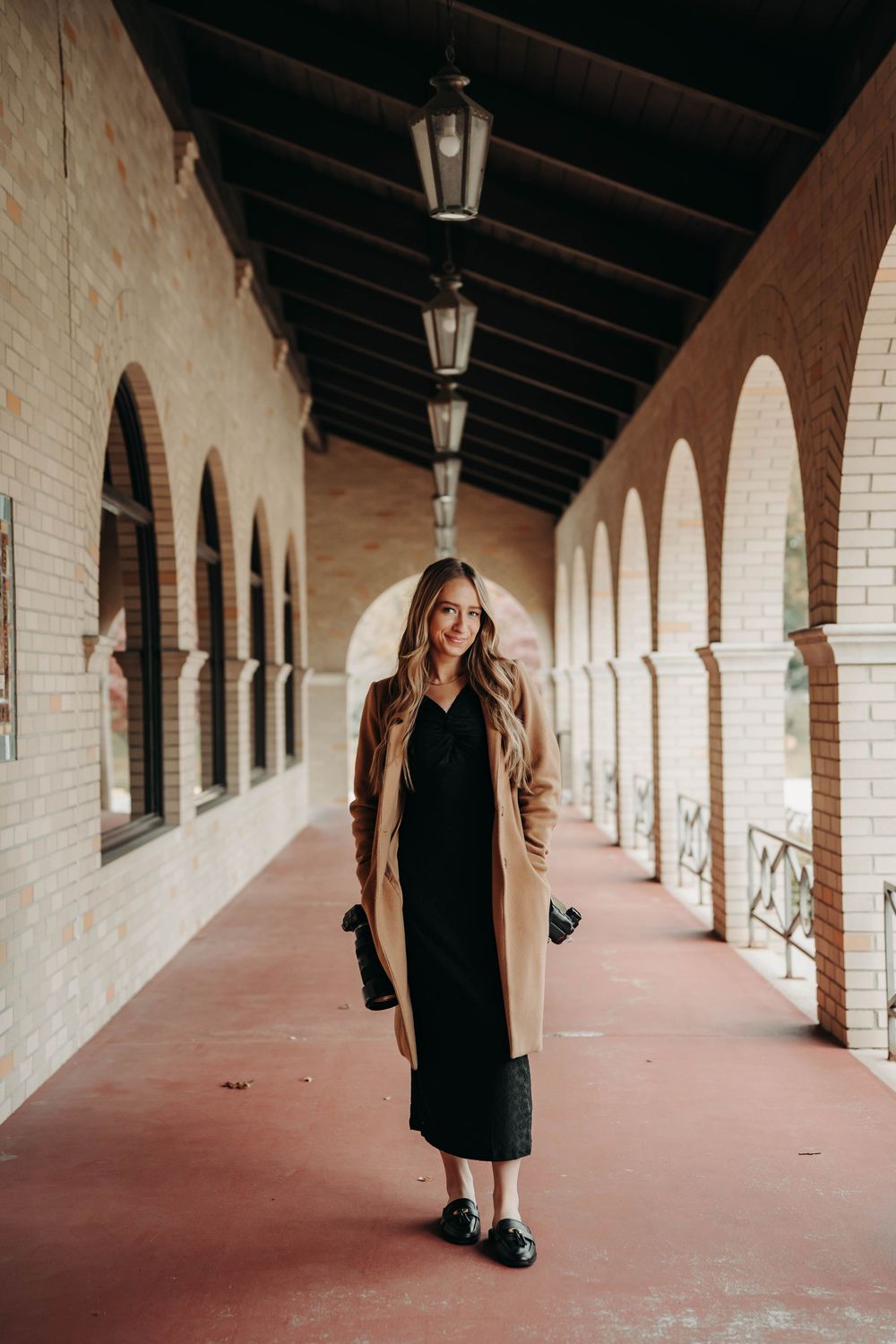 Female wedding photographer in camel coat and black dress posing in an arched brick corridor.