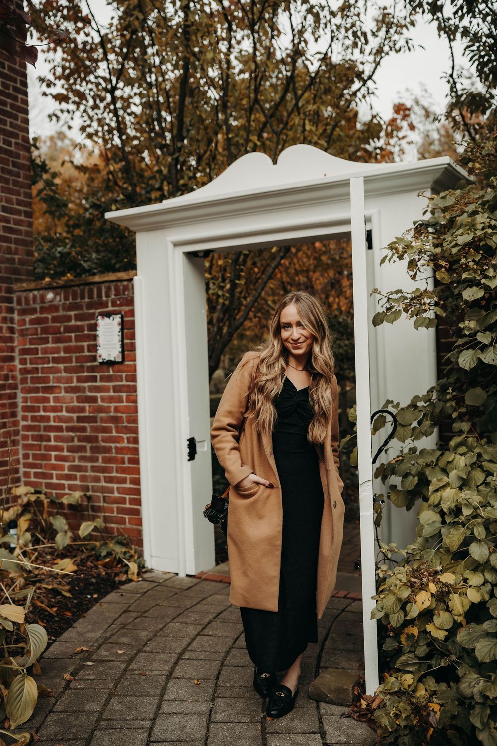 Smiling blonde woman in camel coat and black dress standing in a charming garden doorway in autumn.