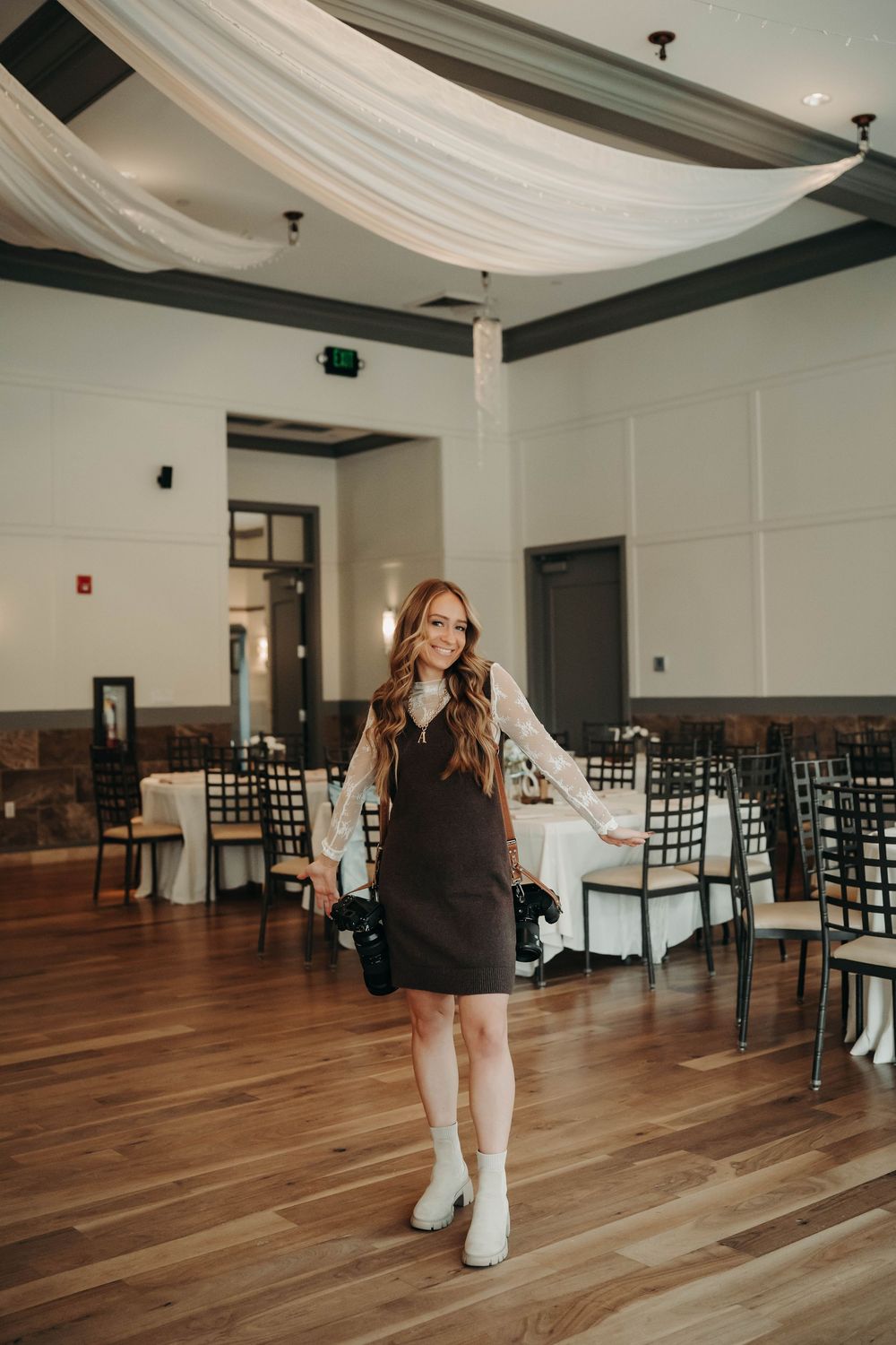 Smiling wedding photographer holding cameras in an elegant ballroom with draped ceilings and dark chairs.