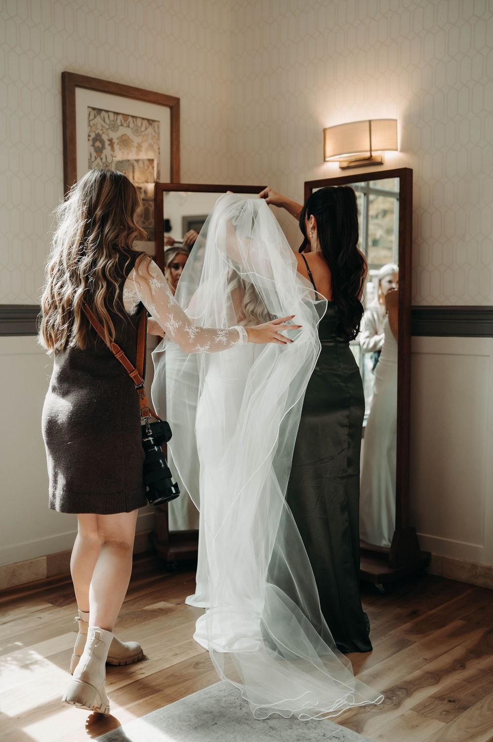 Wedding photographer captures bride in white gown and veil being assisted by bridesmaid before mirror.