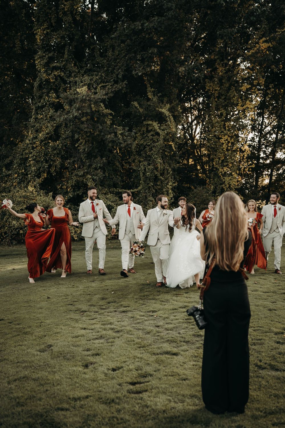 Wedding photographer capturing bridal party in red dresses and tan suits walking outdoors among trees.