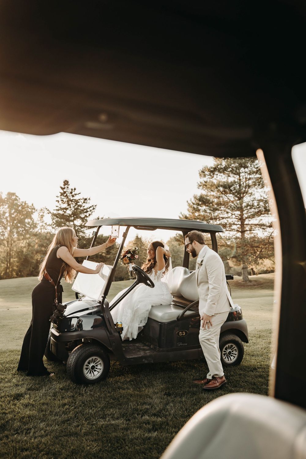 Bride and groom riding a golf cart at sunset with wedding party assistance on a lush green course.