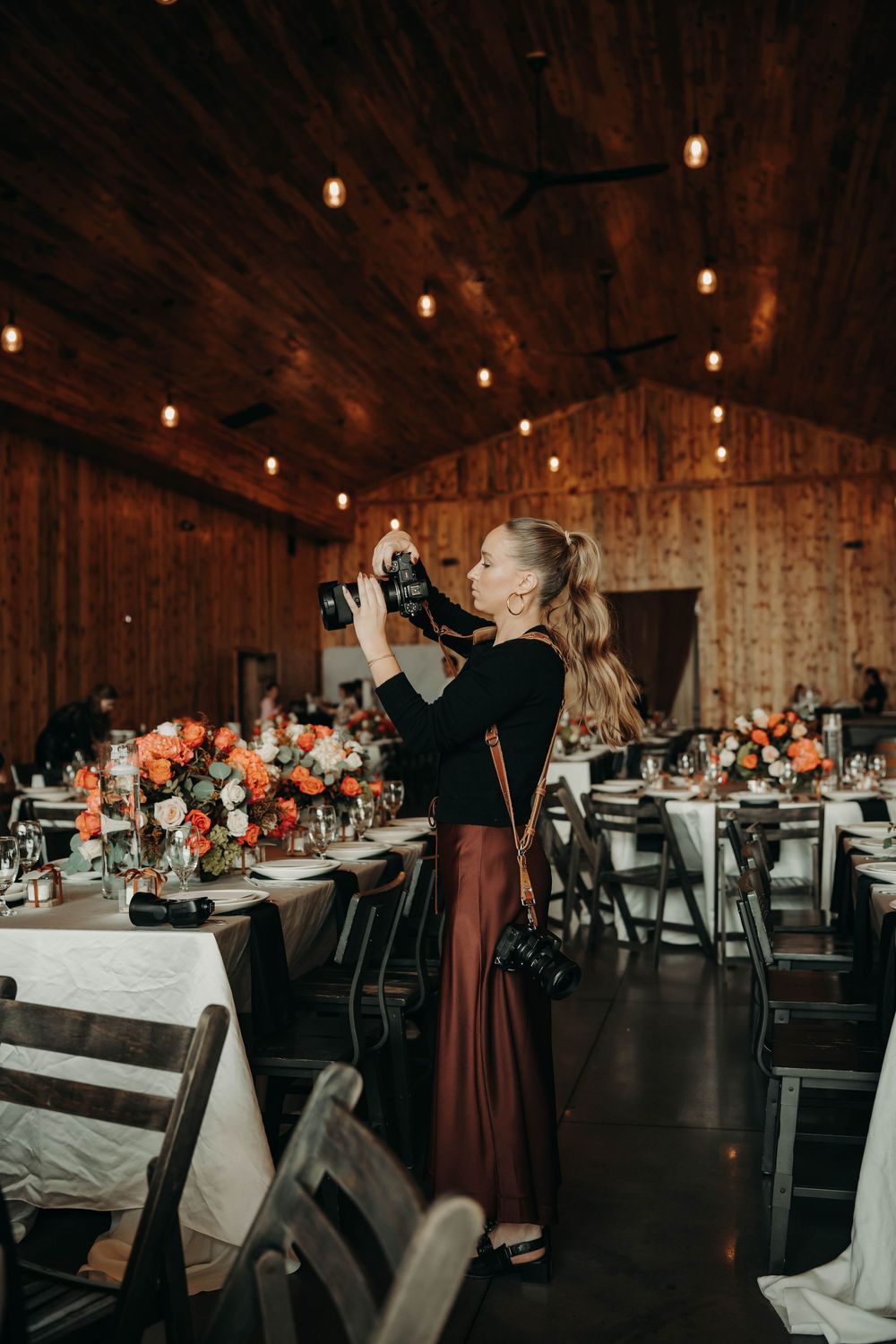Wedding photographer capturing floral centerpieces in a rustic barn reception venue with warm string lights.