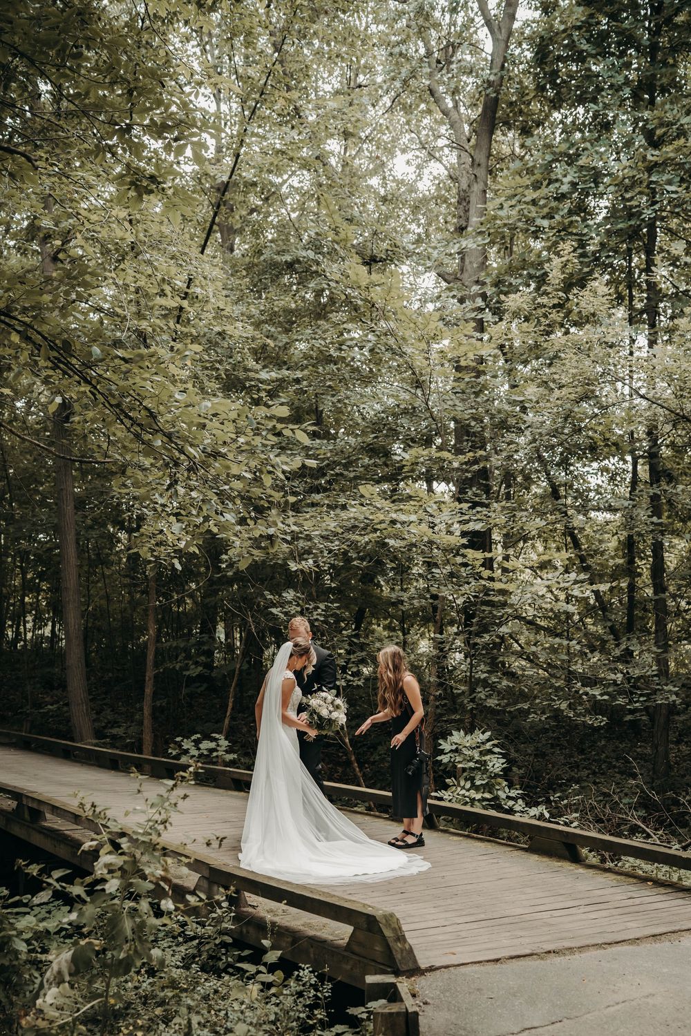 Bride in flowing white wedding gown and veil stands on wooden bridge in lush forest setting.
