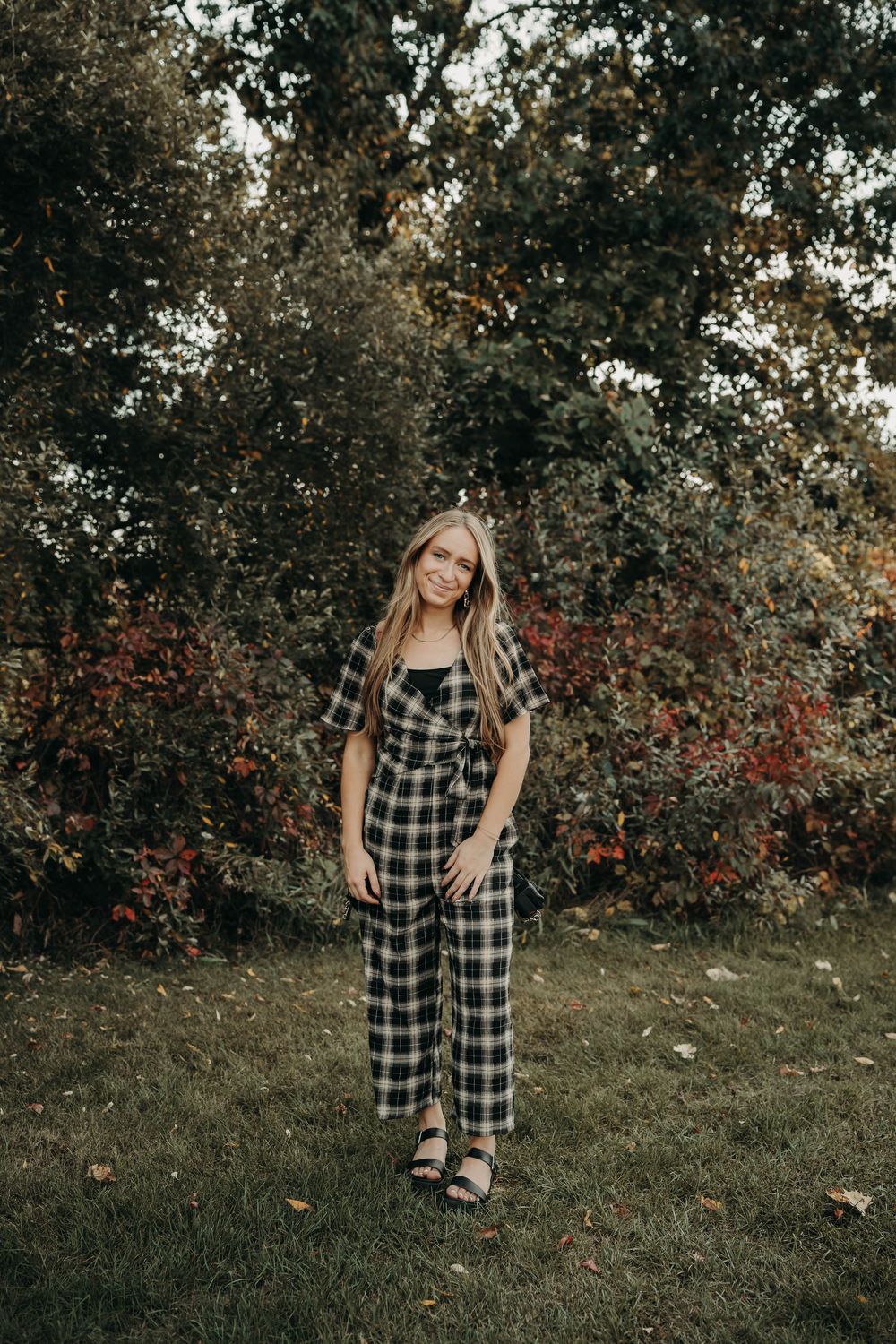 Smiling young woman poses outdoors in plaid jumpsuit against colorful autumn foliage background.