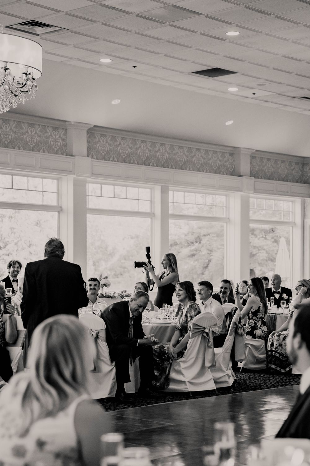Black and white photo of wedding guests seated at reception tables during a toast speech.