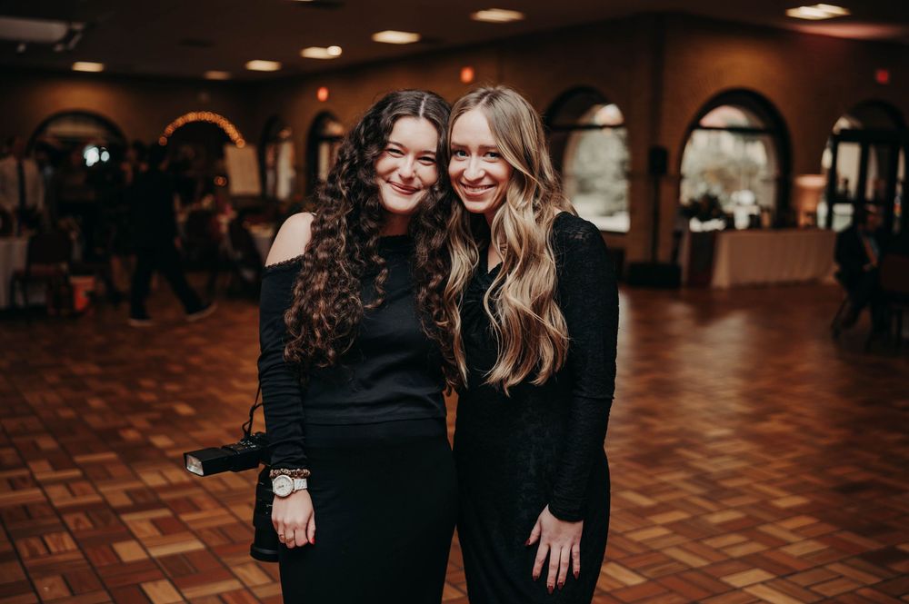 Two female wedding photographers smiling together in black outfits at a warmly lit reception venue.