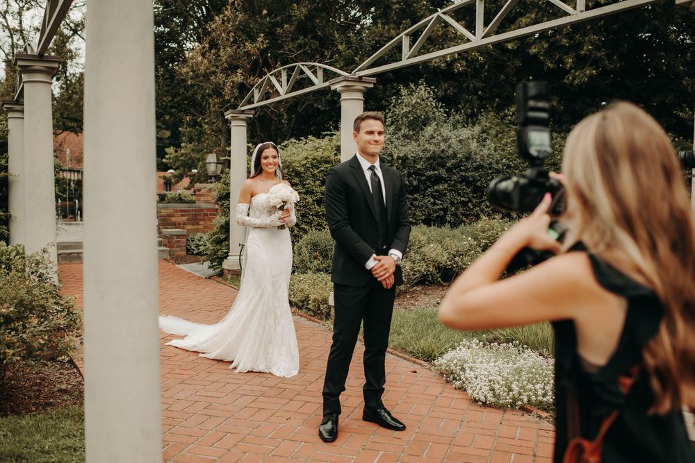 Wedding photographer captures bride and groom portrait under pergola on brick pathway at garden venue.