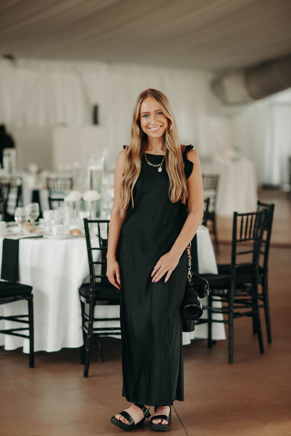 Smiling woman in black maxi dress stands in elegantly decorated wedding reception tent venue.