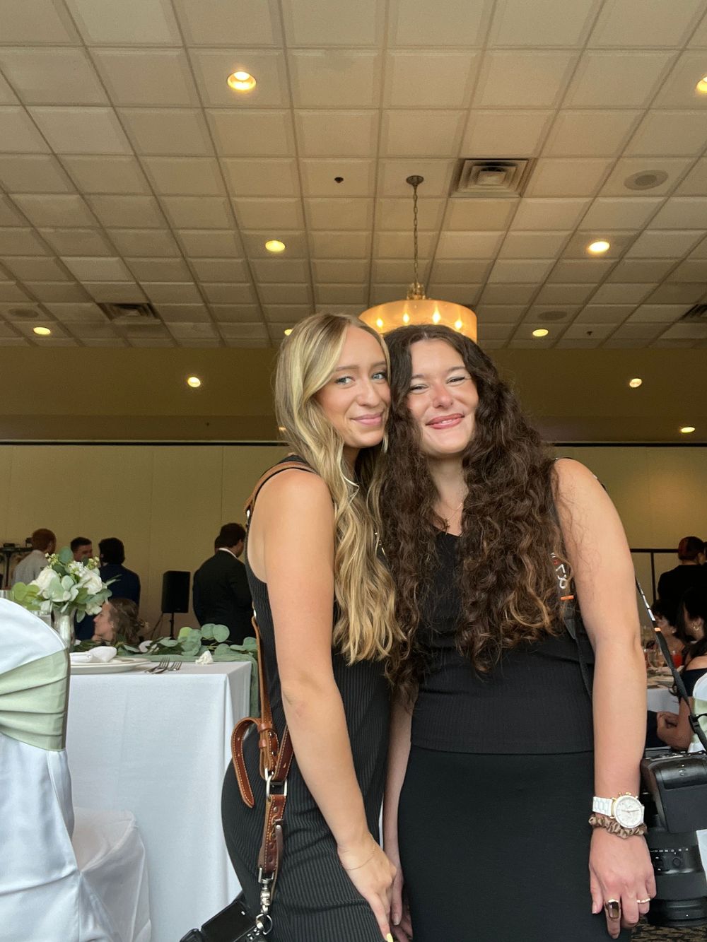 Two women in black dresses smiling together at an elegant wedding reception venue.