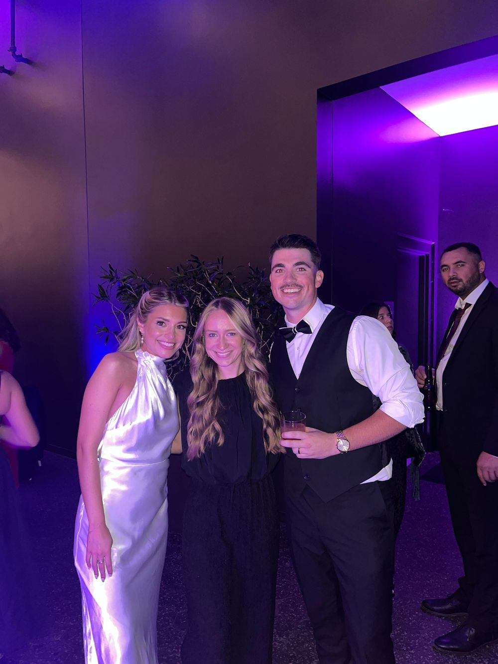 Three guests pose together at a formal wedding reception with purple ambient lighting.