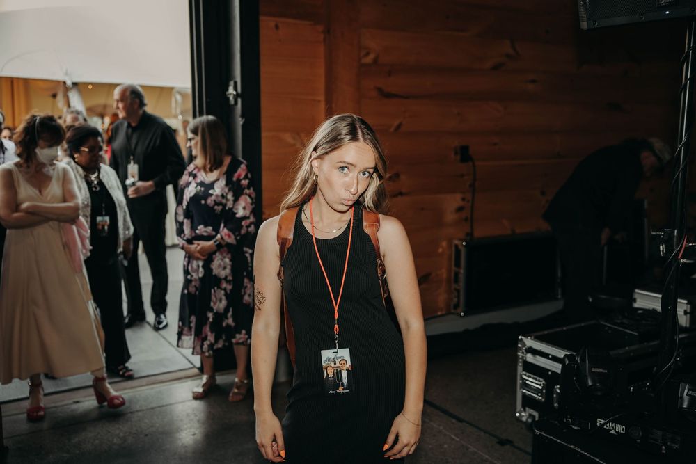 Young woman in black dress wearing event credentials stands inside a rustic wooden wedding venue.