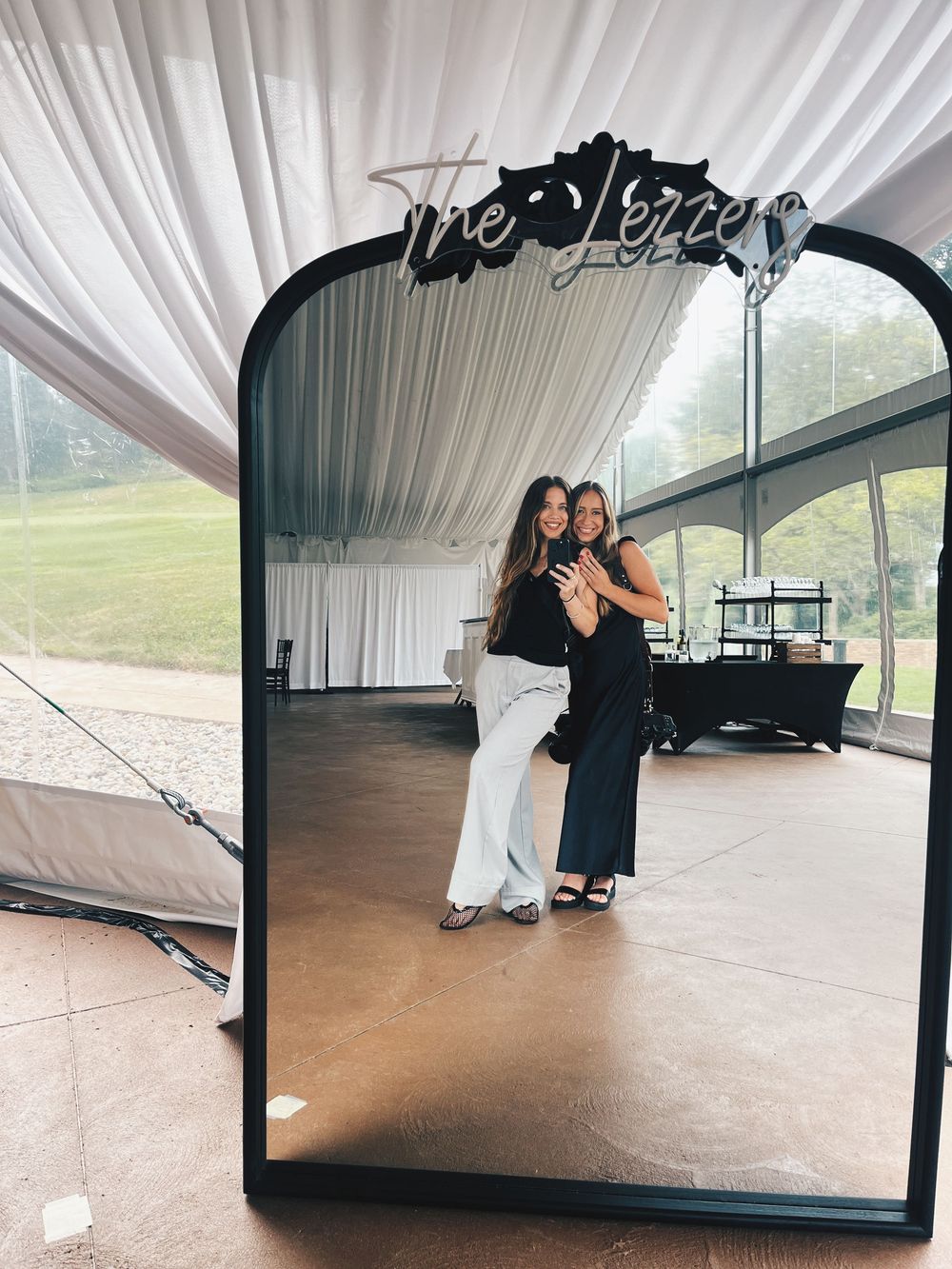 Two women pose together in front of a large decorative mirror at an elegant wedding reception venue.