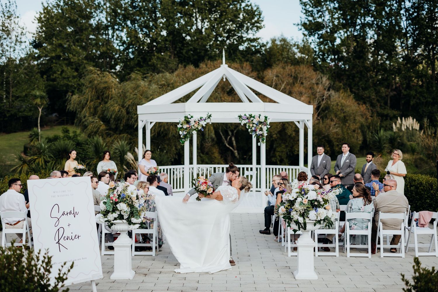 A beautiful outdoor wedding ceremony under a white gazebo surrounded by guests in a garden setting.