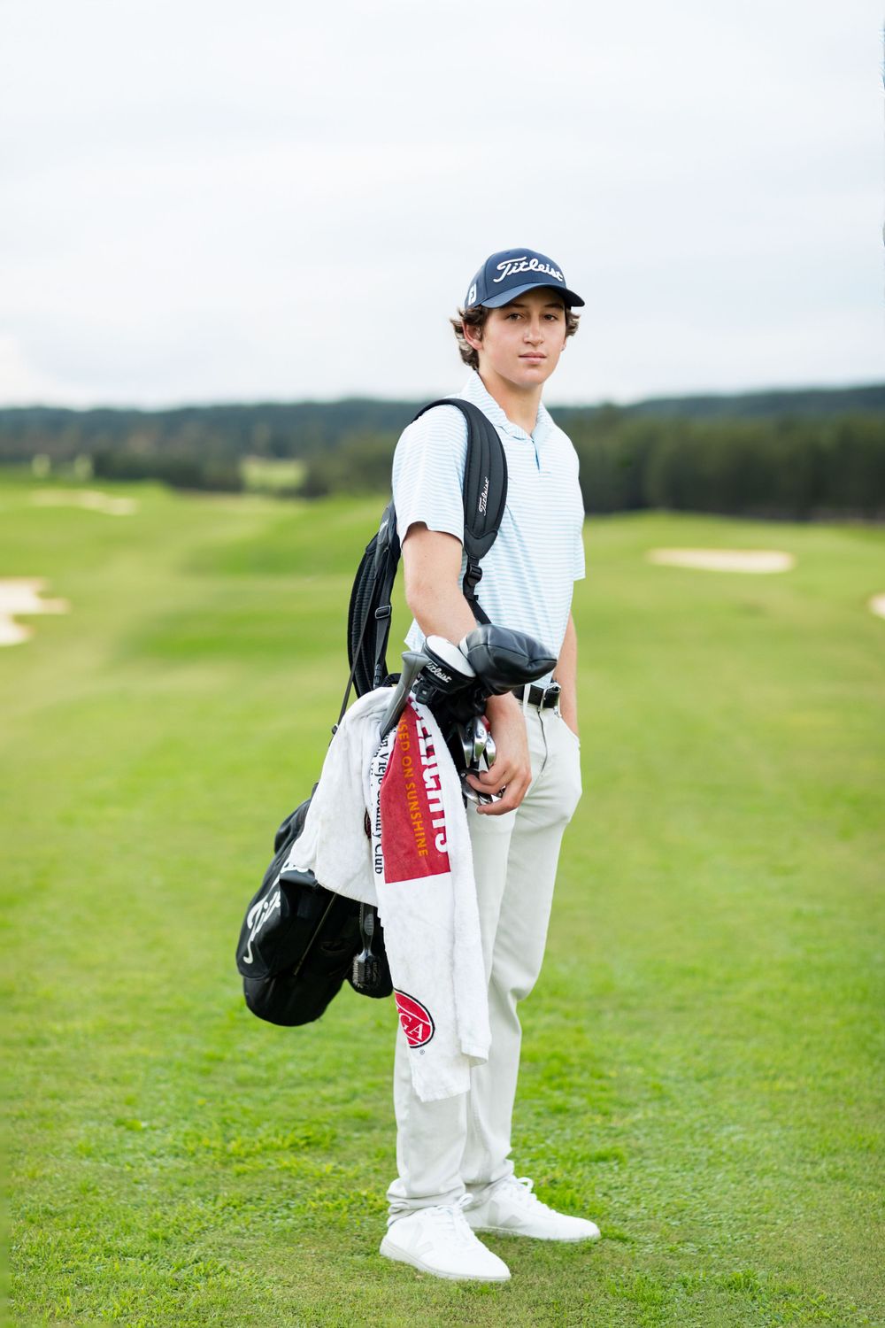Young golfer in white attire stands on golf course carrying clubs and wearing a baseball cap.
