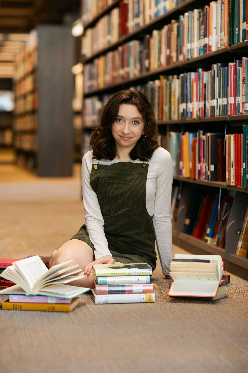 Student wearing a green corduroy jumper sits on library floor surrounded by open books and bookshelves.