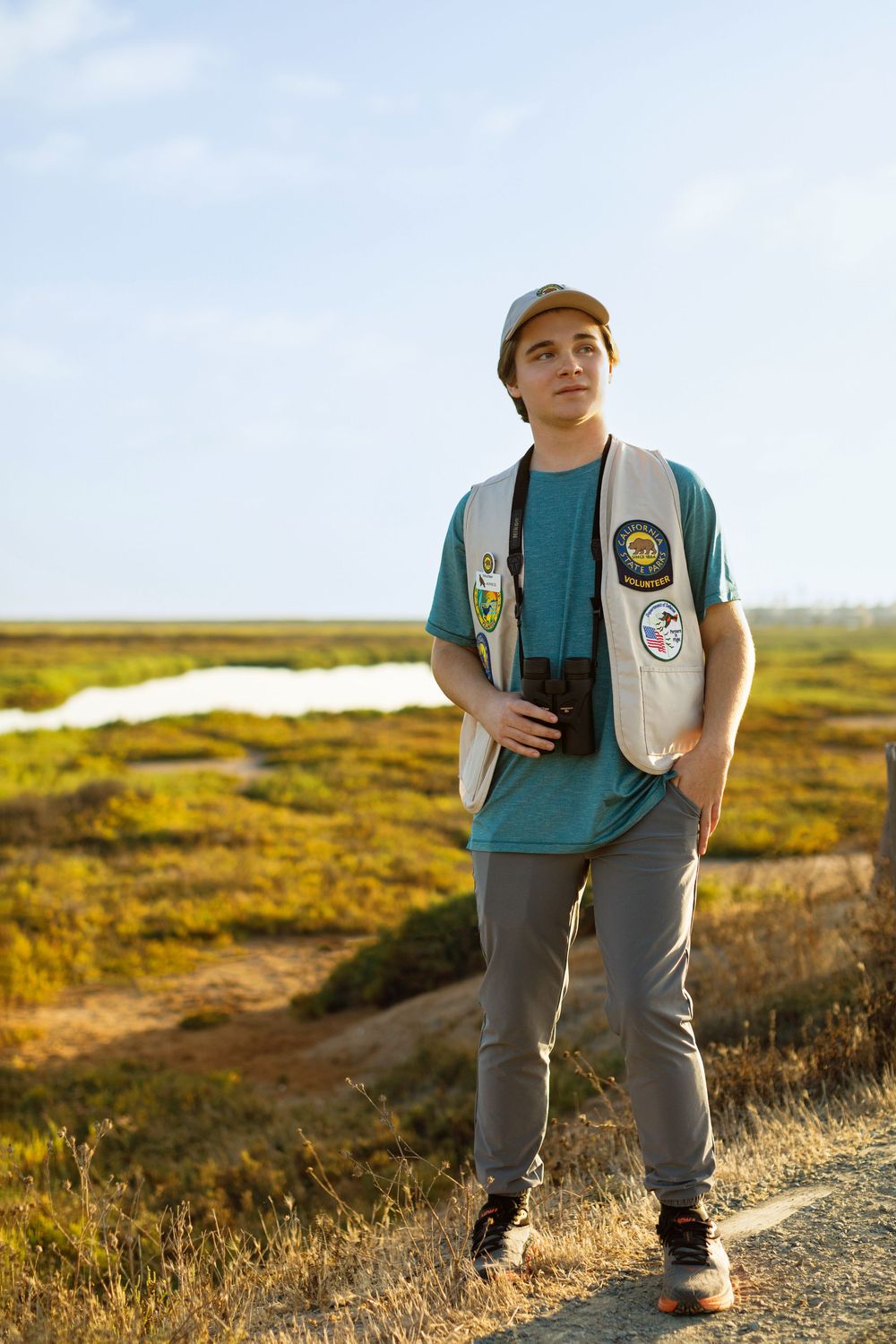A person wearing a vest with badges stands on a hiking trail during golden hour in a grassy coastal landscape.
