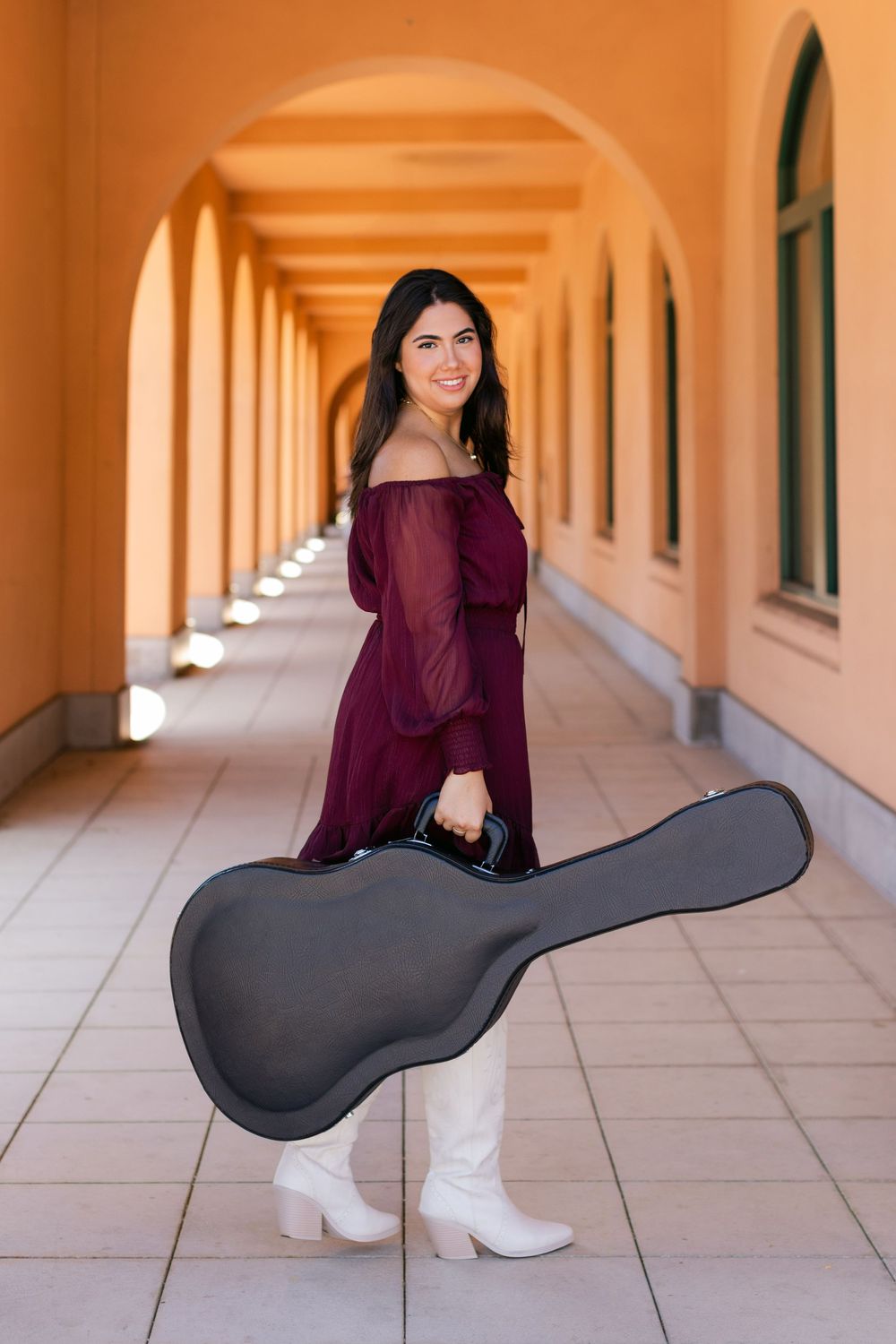 A person wearing a burgundy dress and white boots holds a guitar case in an arched hallway.