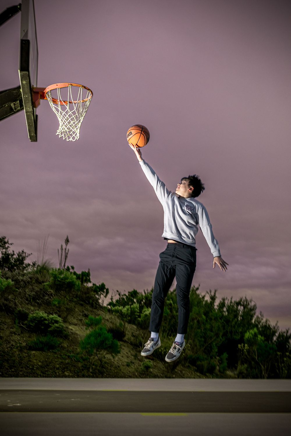 Basketball player leaping high to dunk on an outdoor court at dusk with dramatic purple sky in background.