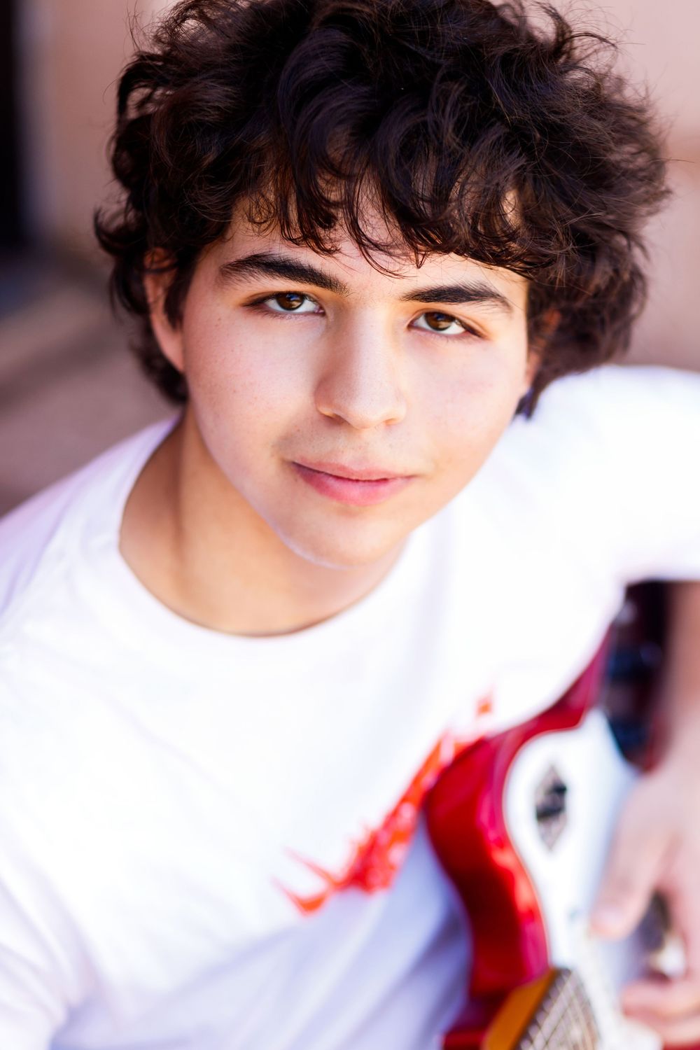 Young teen boy with curly dark hair holding a red electric guitar, wearing a white t-shirt, looking at camera.