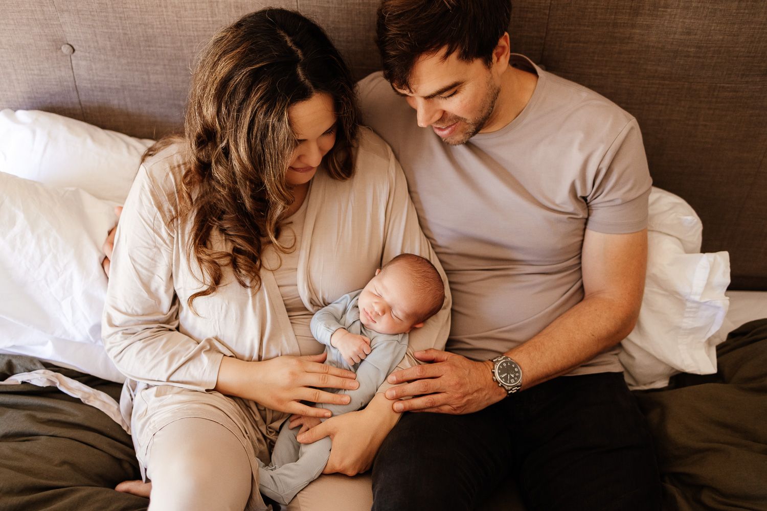A couple sitting on a bed, lovingly gazing at their newborn baby wrapped in a blue onesie.
