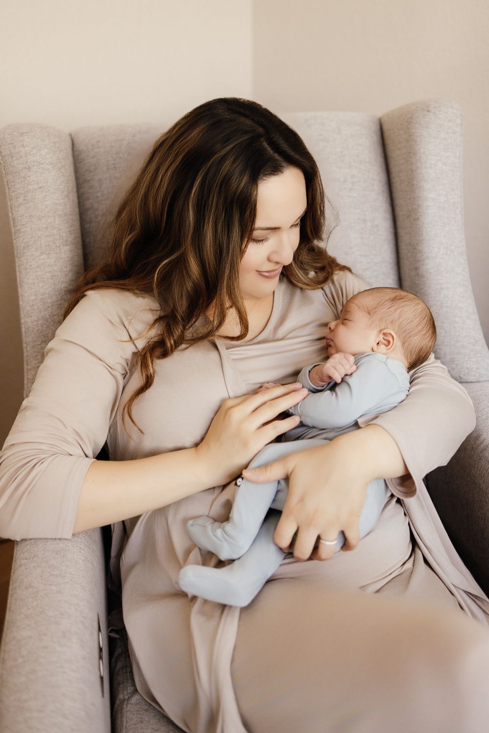 A mother in a beige dress sits in a gray chair, lovingly cradling her newborn baby dressed in blue.