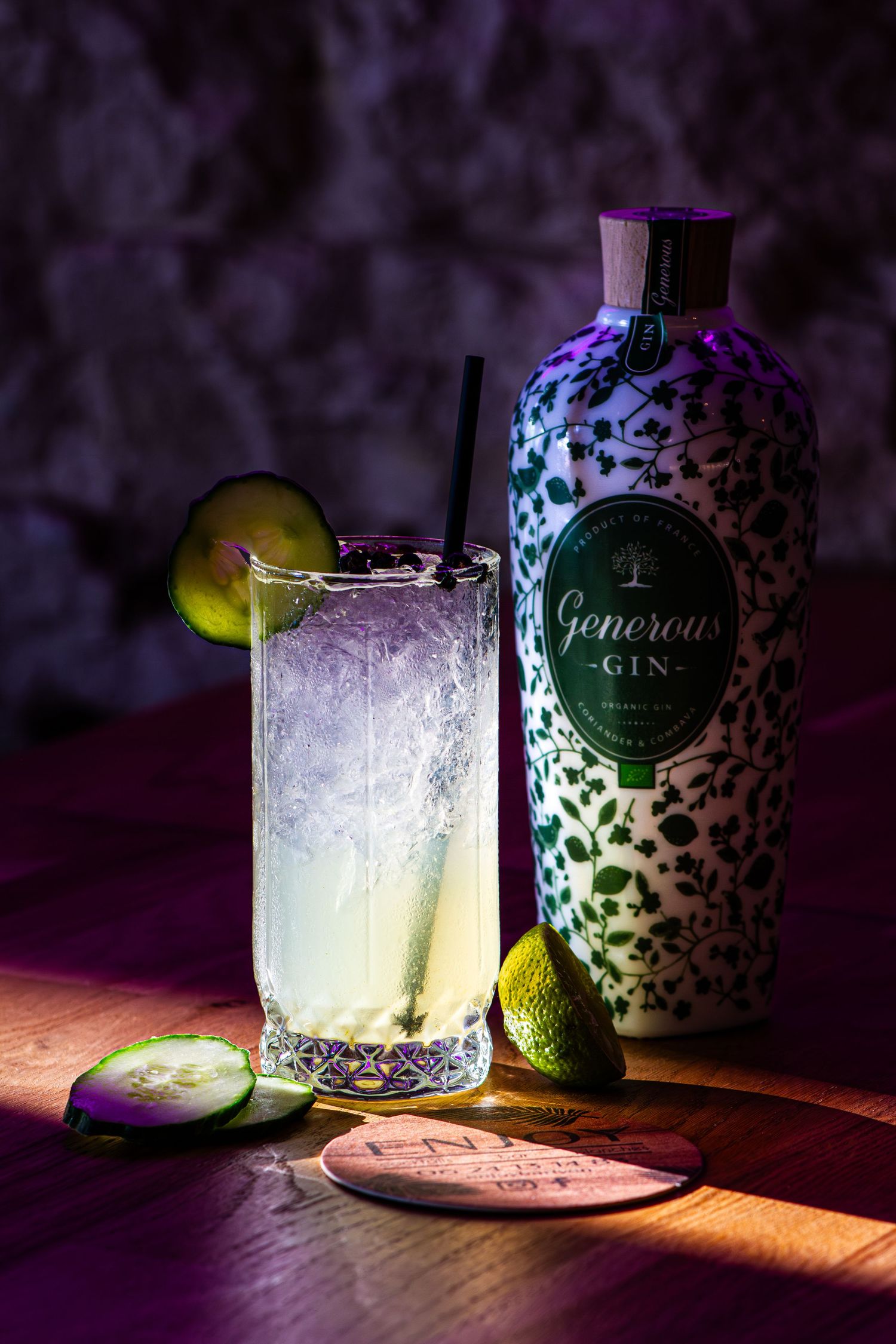 Generous Gin bottle beside a tall cocktail glass with ice, cucumber garnish, and a straw on a wooden bar.