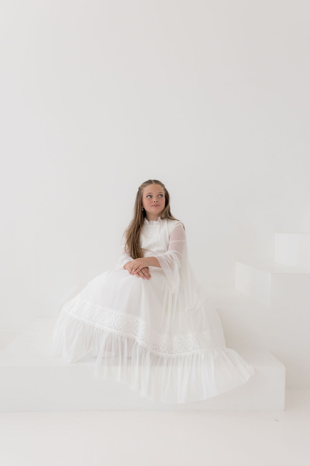 Young girl in elegant white long-sleeve tulle dress seated against white background, perfect for formal occasions.