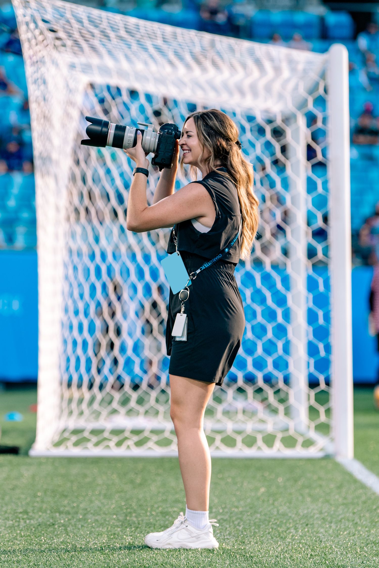 Female sports photographer smiling while shooting with a telephoto lens at a soccer stadium.