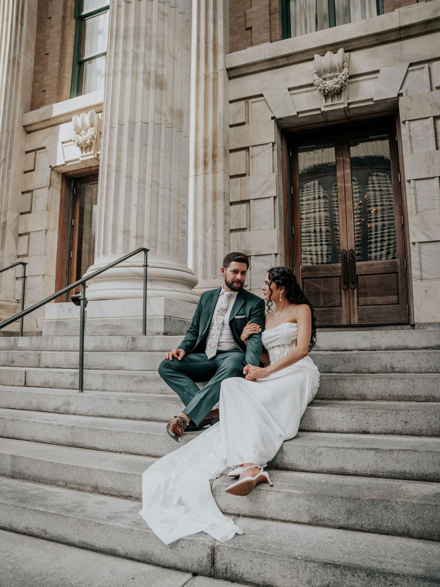Bride and groom sitting on stone steps of elegant building with columns, sharing a romantic moment in wedding attire.