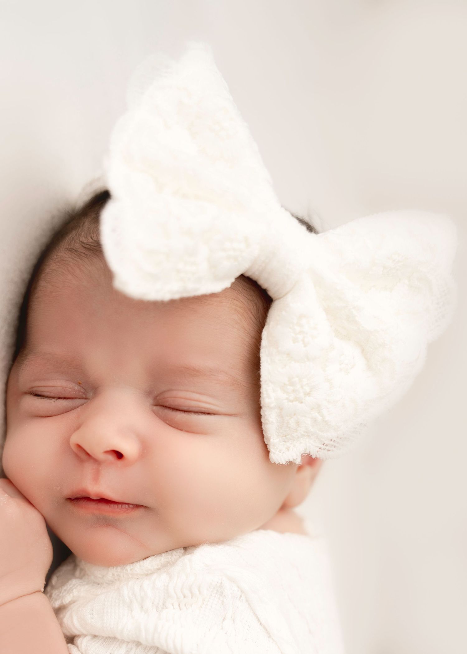 A sleeping baby wearing a white bow headband rests peacefully against a soft white background.