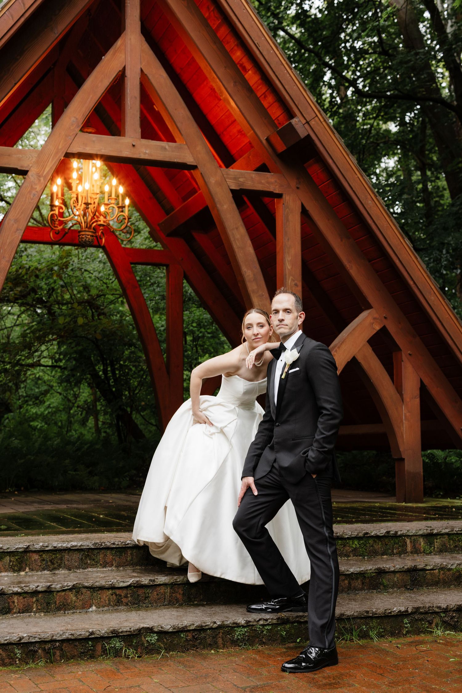 Wedding couple posing under dramatic red wooden arch structure surrounded by lush green trees.