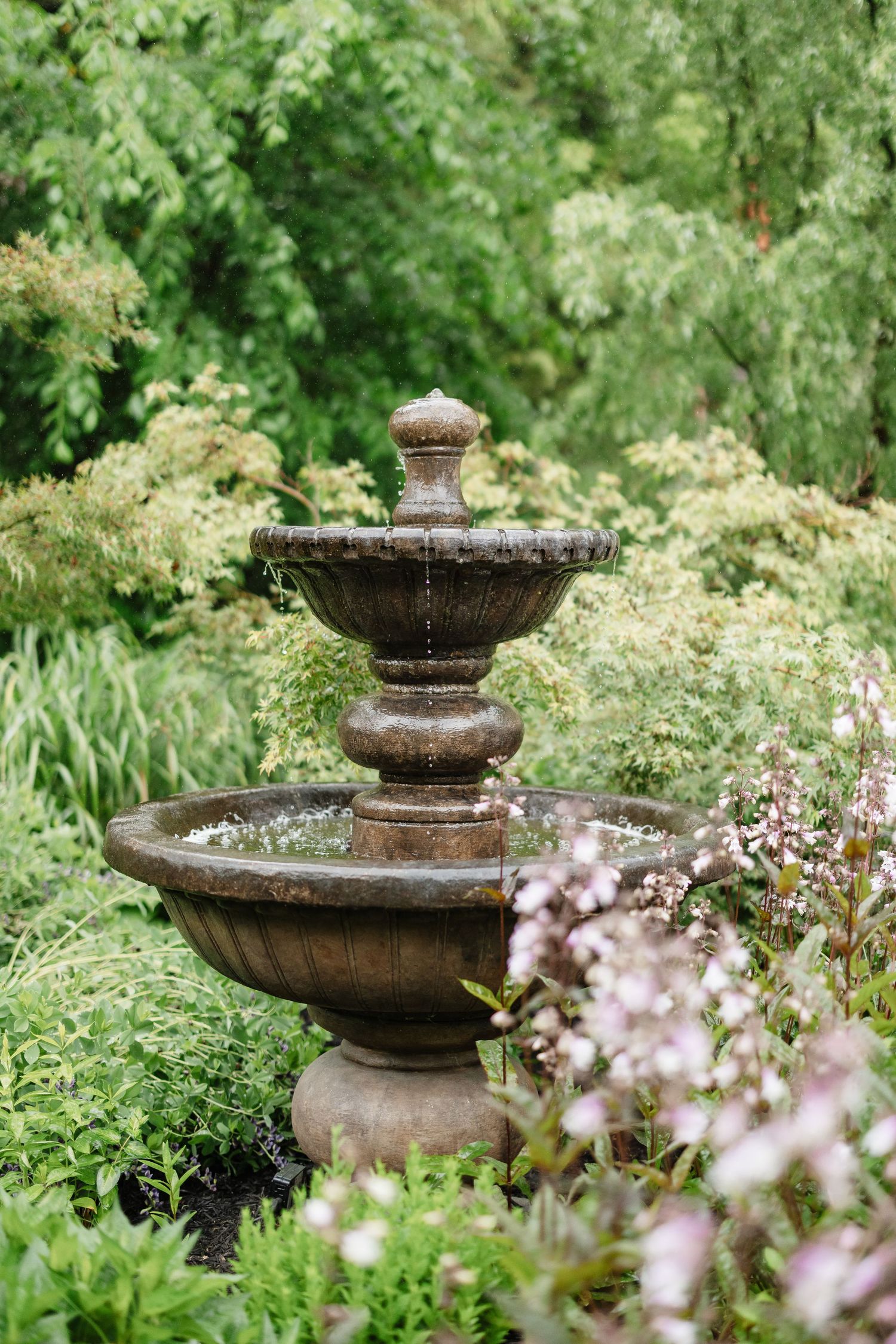 A classic three-tiered stone garden fountain surrounded by lush greenery and pink flowering plants.