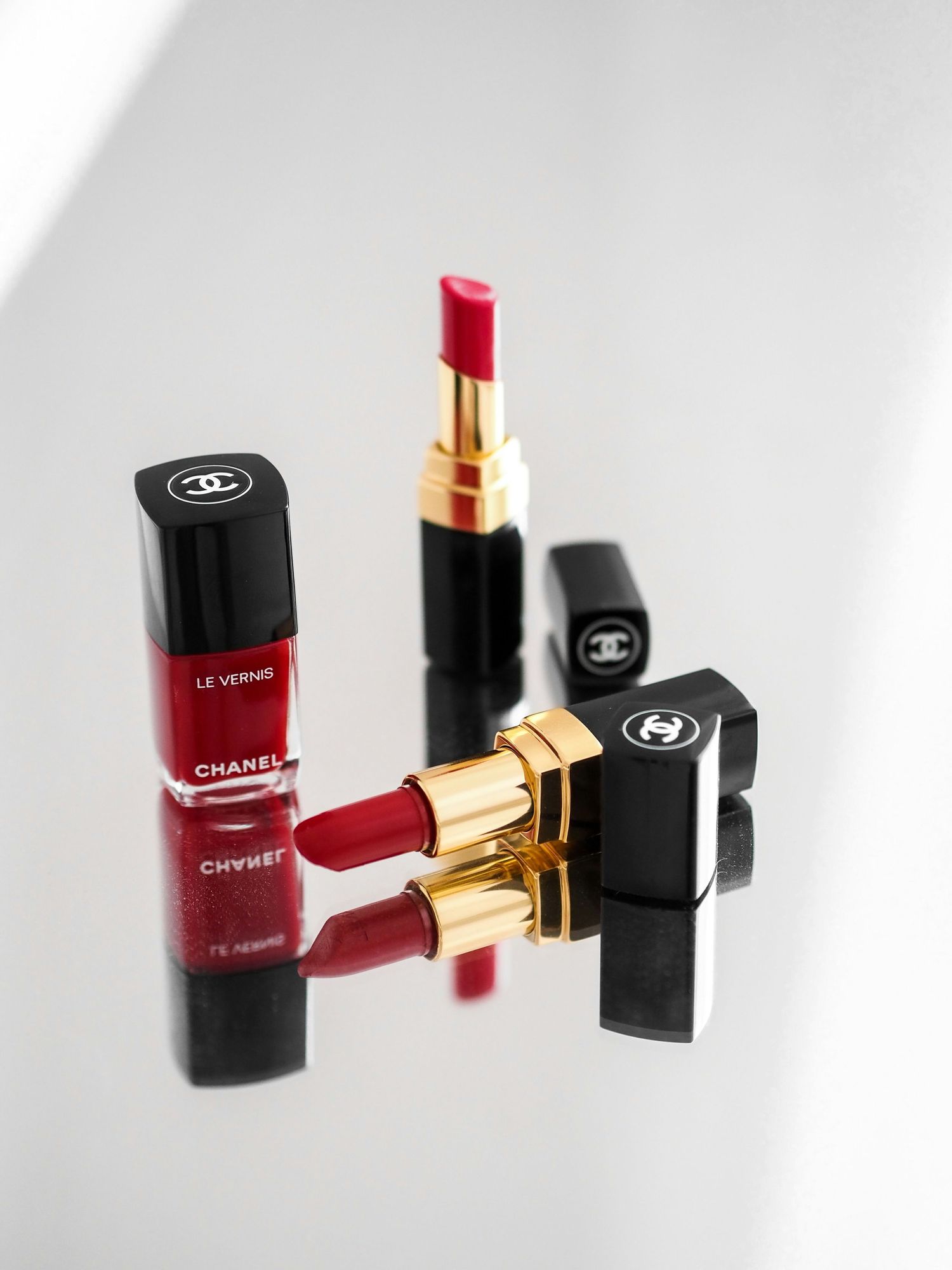 Chanel Le Vernis red nail polish and Rouge Coco lipsticks in black and gold cases on a reflective white surface.