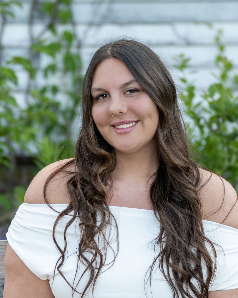 Young woman with long dark wavy hair smiling outdoors, wearing a white off-shoulder top with green foliage background.