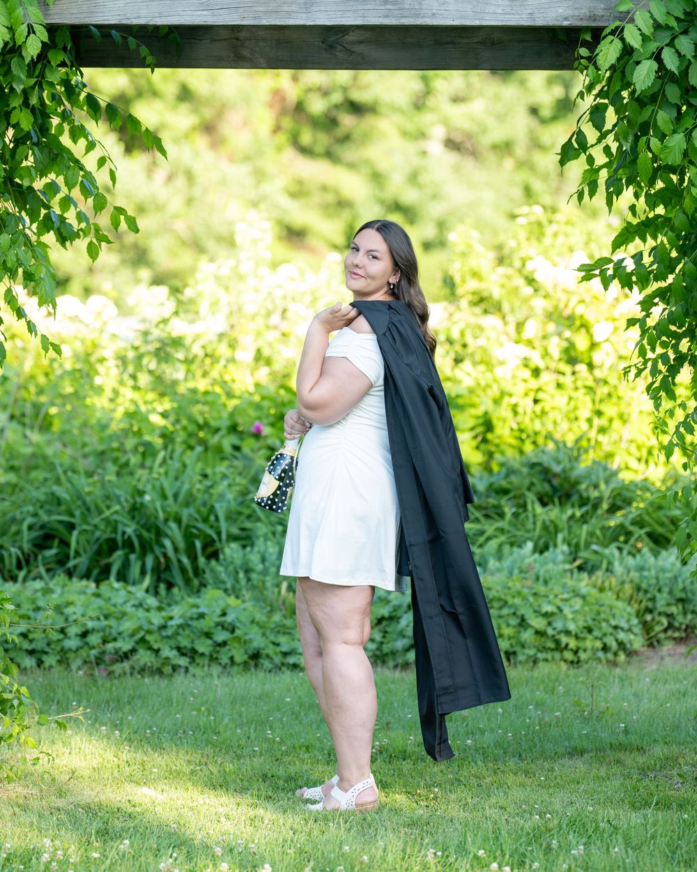 Woman in white dress holding black graduation gown poses in lush green garden setting.