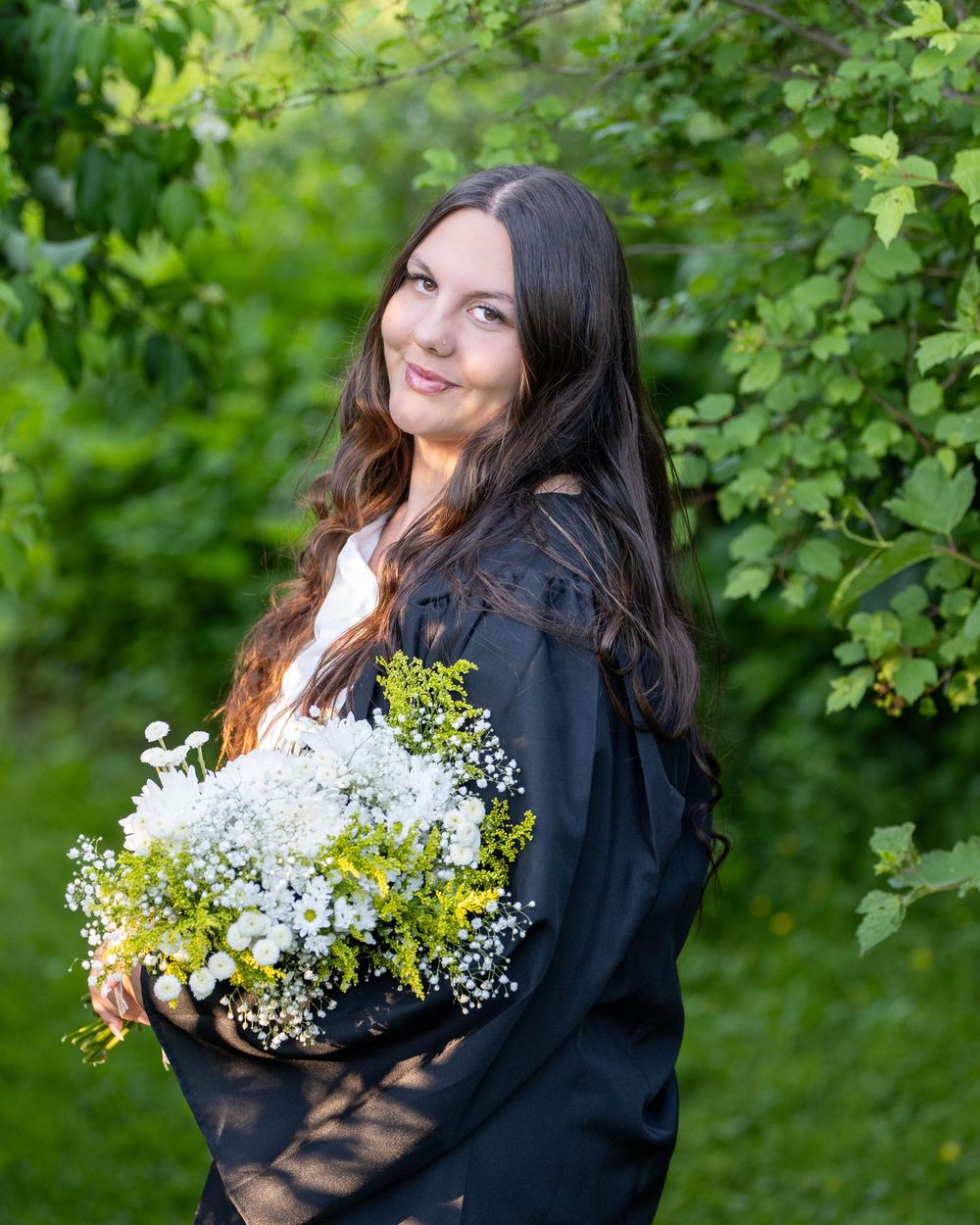 Young woman in graduation gown holding white wildflower bouquet, smiling outdoors among lush green trees.