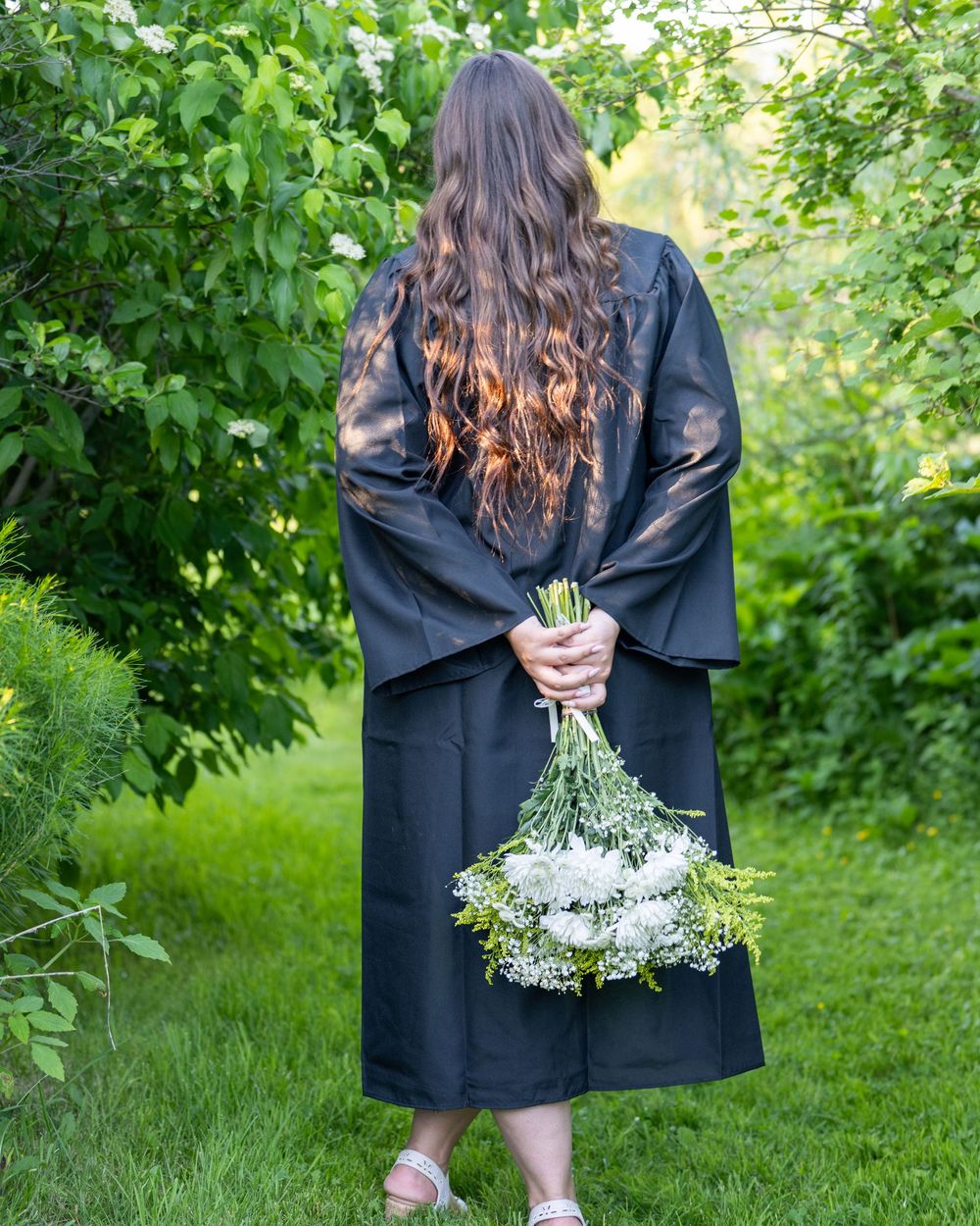 Graduate in black gown holding white flower bouquet, standing in lush green garden, viewed from behind.