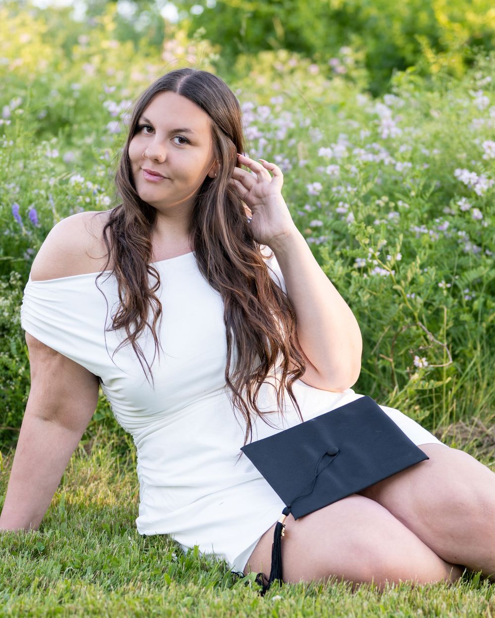 Young woman in white off-shoulder dress holding graduation cap while sitting in a flower field.