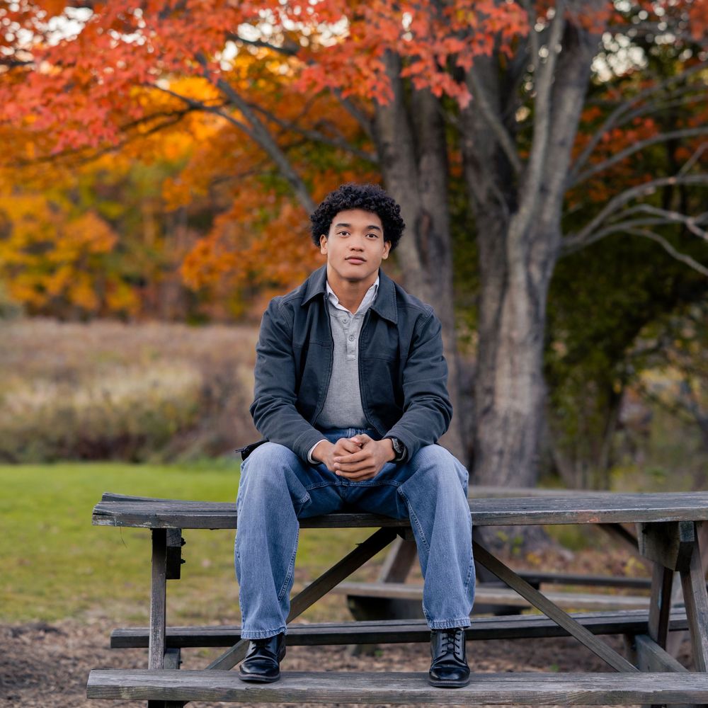 Young man sitting on a picnic table in a park with vibrant autumn foliage in the background.