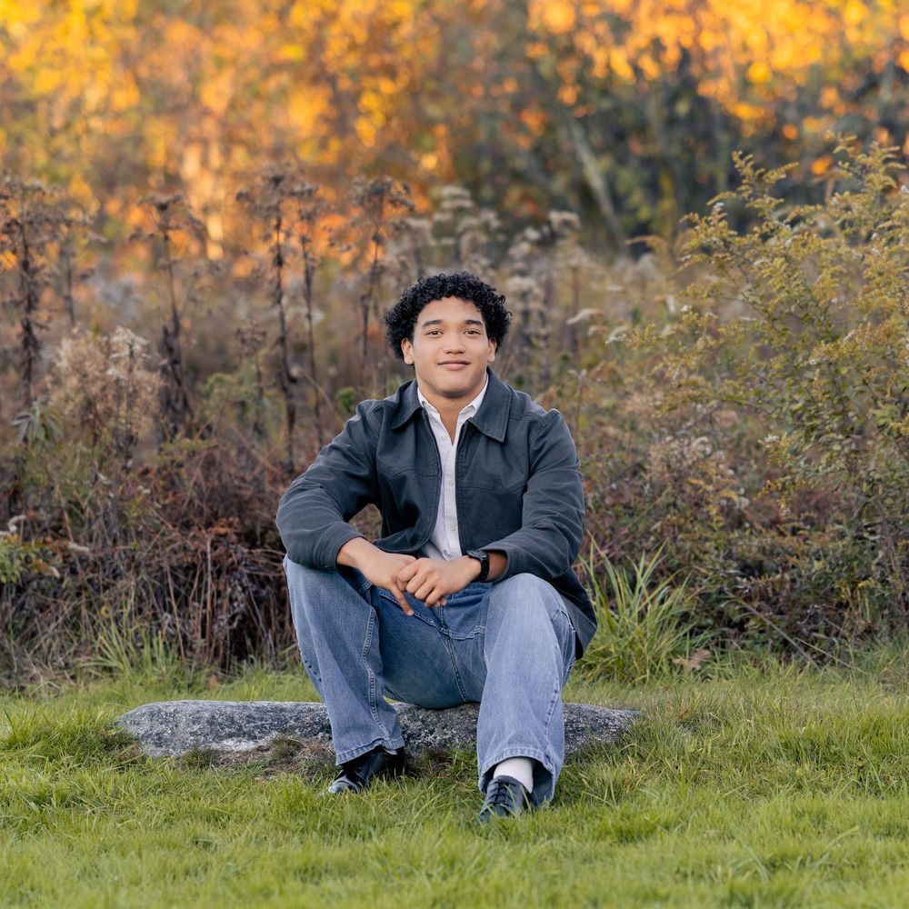 Young man sitting on a rock in a field with colorful autumn foliage in the background.