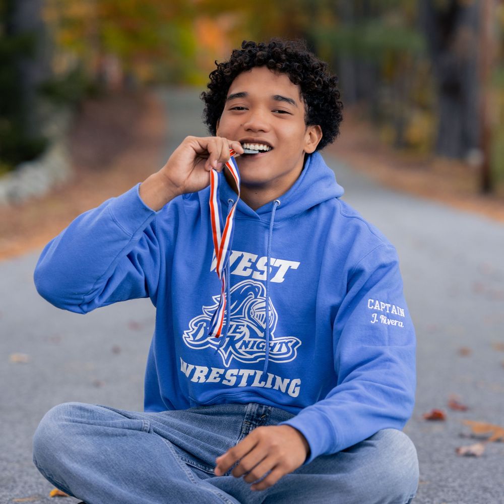 Young athlete in blue West Knights Wrestling hoodie biting a medal while sitting cross-legged outdoors.