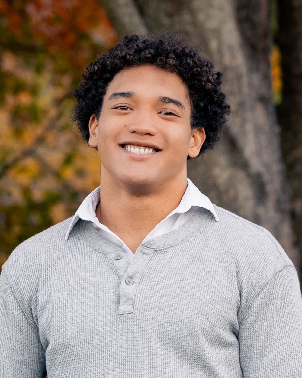 Smiling young man with curly hair wearing a gray waffle-knit polo shirt outdoors with autumn foliage background.