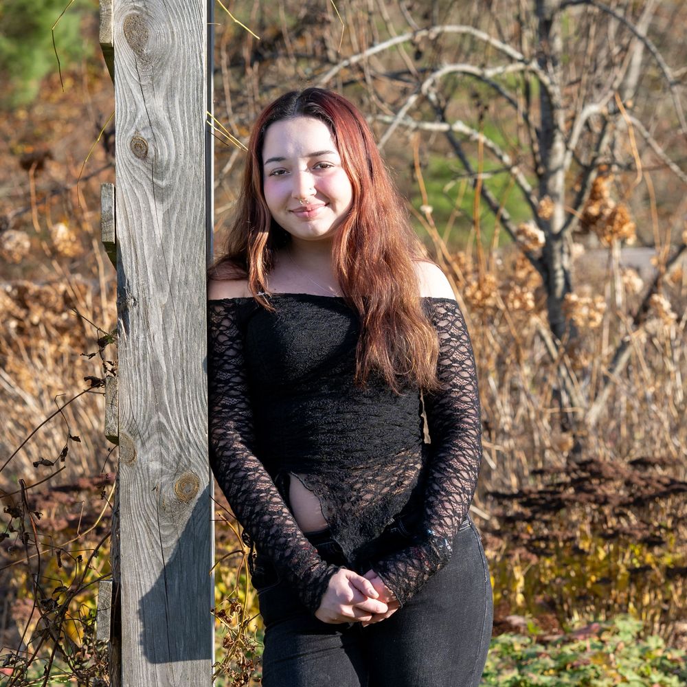 Young woman with long red hair wearing a black off-shoulder top, posing outdoors near a wooden post in autumn.