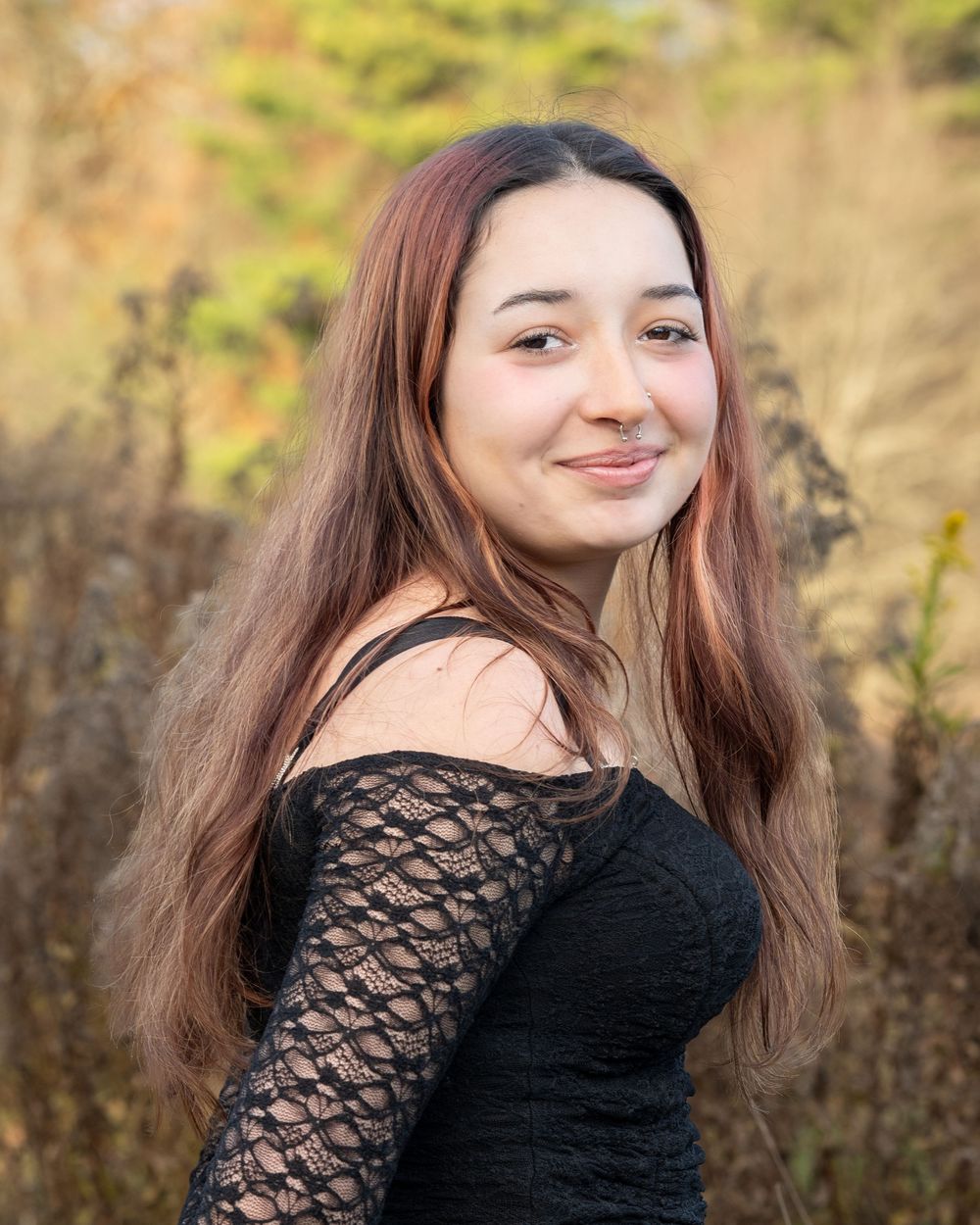 Young woman with long auburn hair smiling outdoors in a black lace off-shoulder top with autumn foliage background.