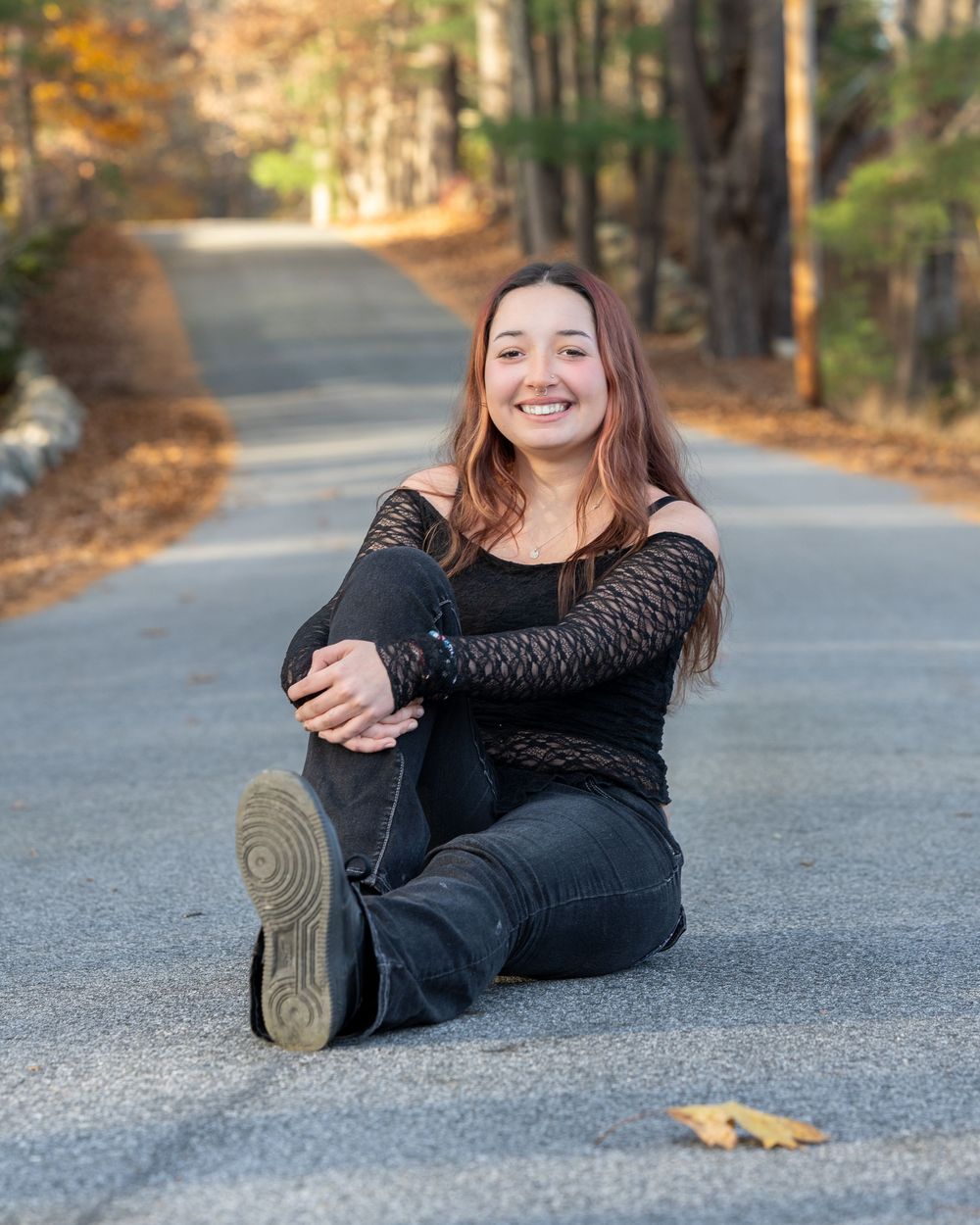 Young woman in black outfit sitting on a tree-lined road, smiling, wearing dark jeans and boots in autumn setting.
