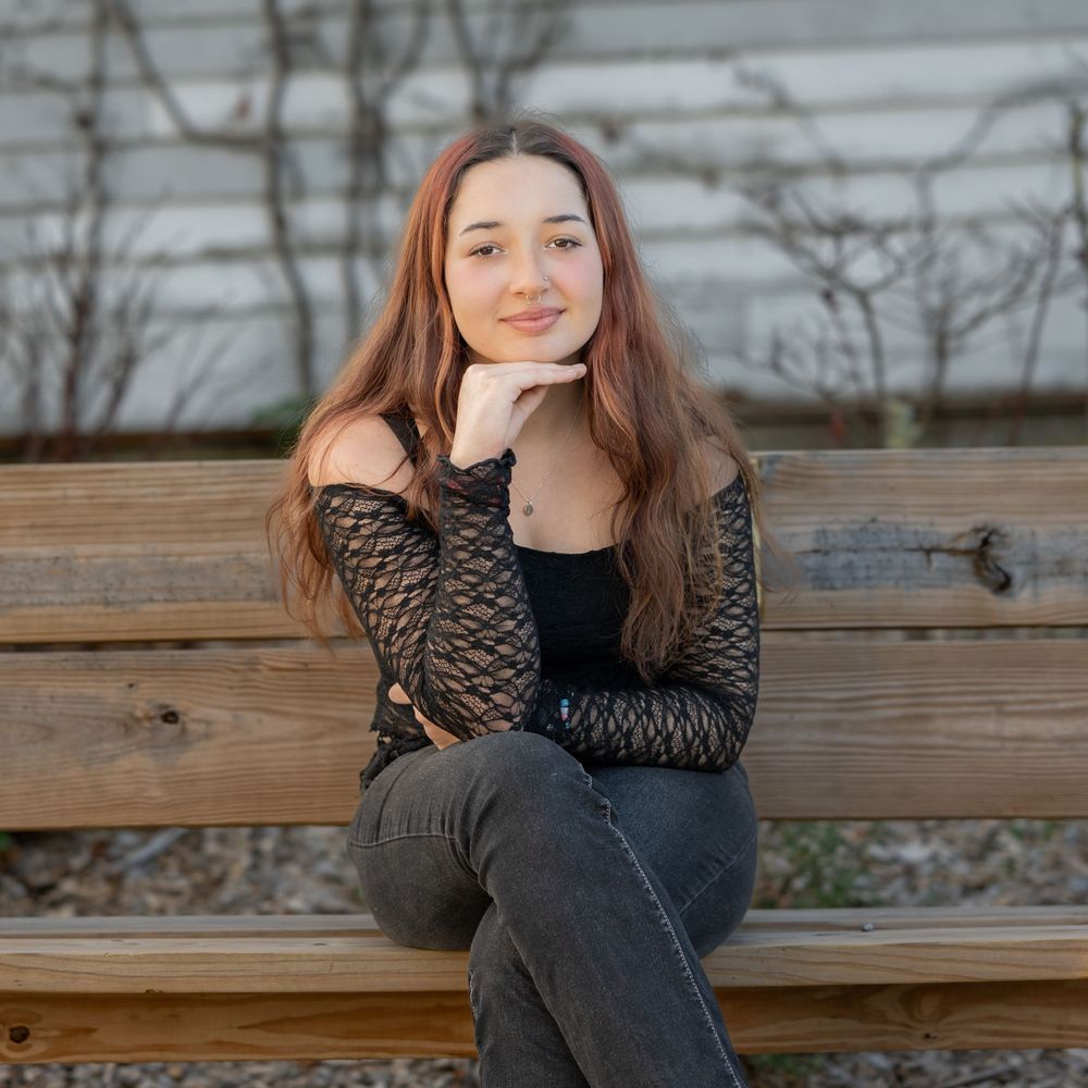 Young woman with long hair sitting on a wooden bench outdoors, wearing a black top and gray jeans.